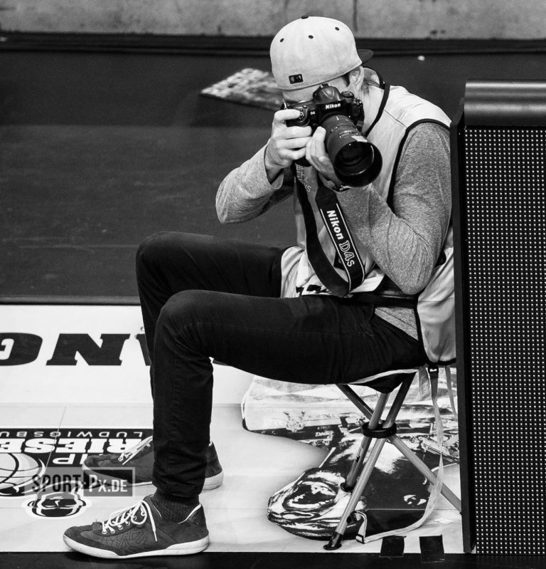 Ein Fotograf sitzt auf einem Stuhl und hält seine Kamera Richtung Basketball Feld gerichtet.